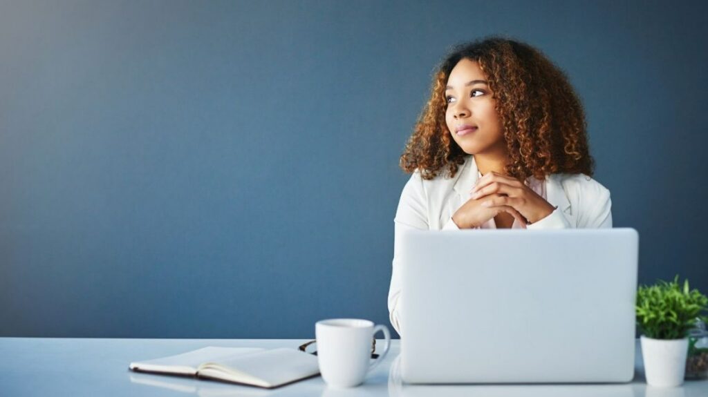 A woman is looking to her right while thinking, with a laptop and a mug on her desk.