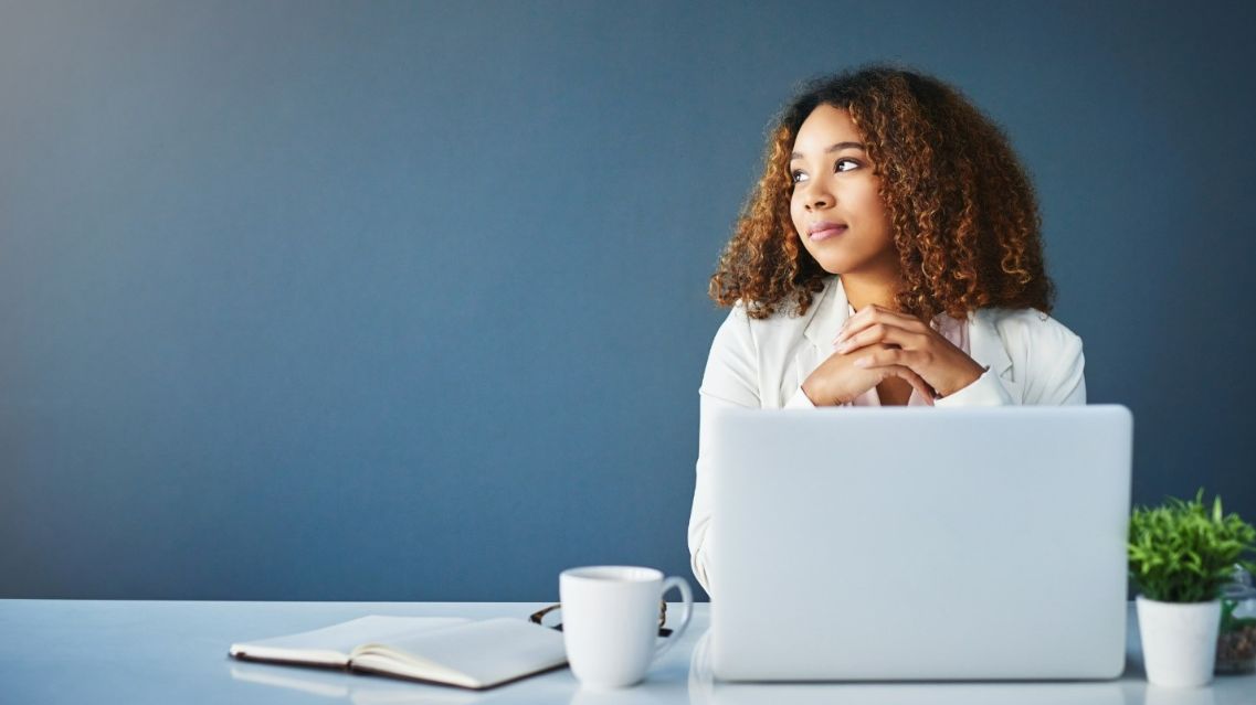 A woman is looking to her right while thinking, with a laptop and a mug on her desk.